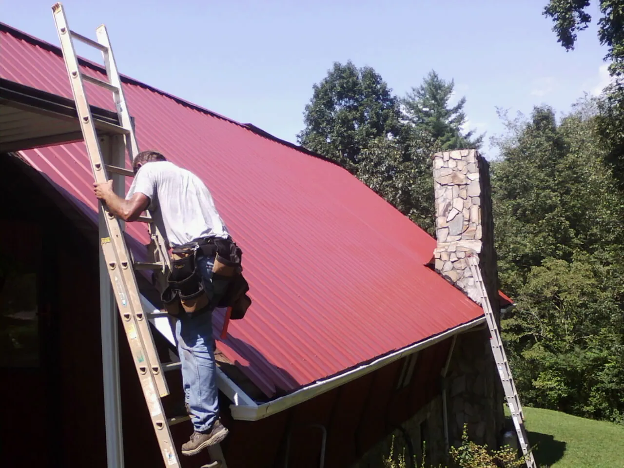 Roofer on a ladder beside a red metal roof and stone chimney — Soffit & Fascia Repair by Roofing Brothers in Randlett